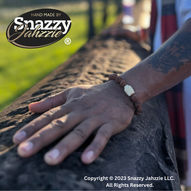 A close-up of a hand wearing a 9mm Buddha Vajra Bodhi Rudraksha Bracelet with brown and white beads resting on a stone surface. The background is blurred, showing an outdoor setting perfect for meditation. The top left corner features the logo "Hand Made by Snazzy Jahzzie." The bottom text reads "Copyright © 2023 Snazzy Jahzzie LLC. All Rights Reserved.