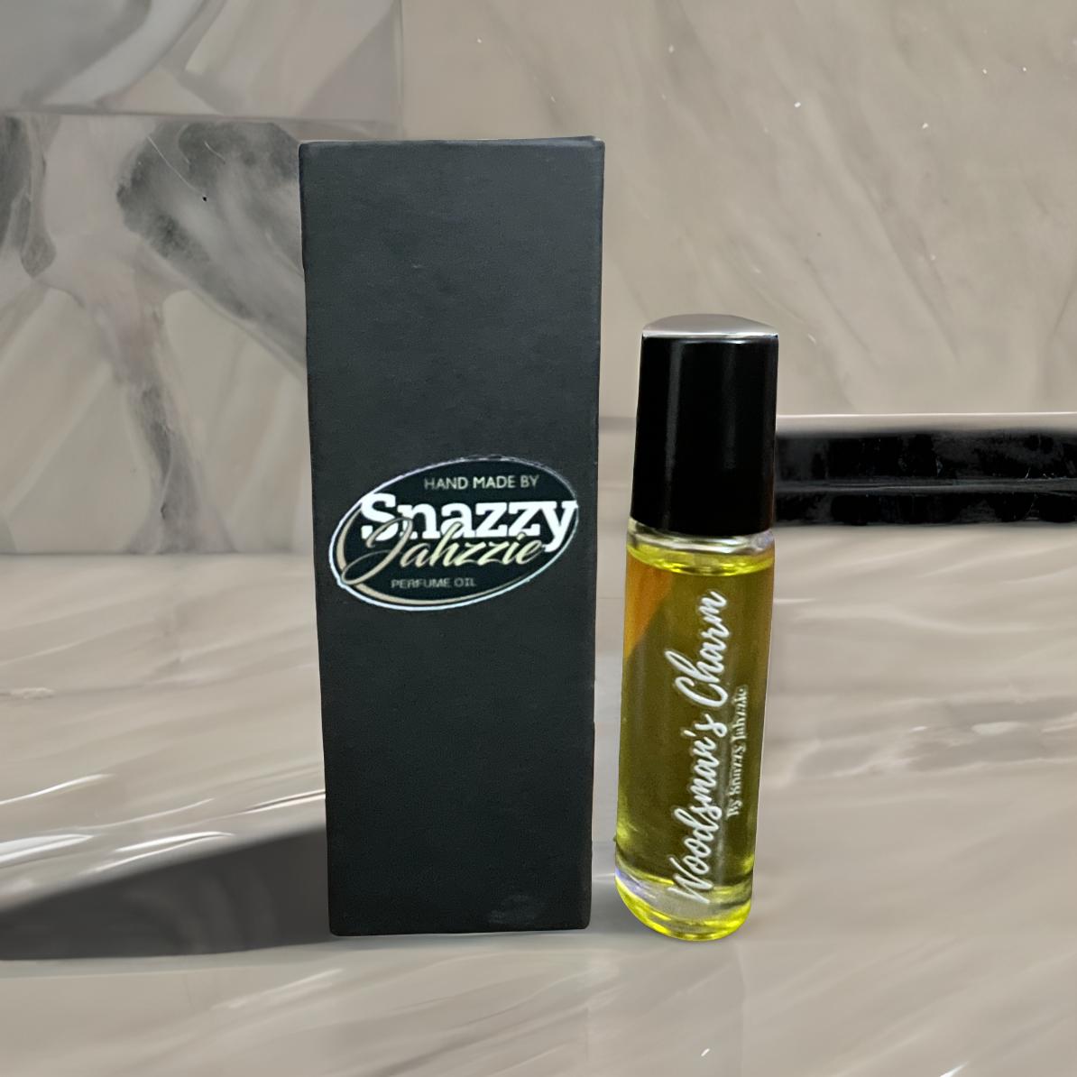 A small bottle of Woodsman's Charm By Snazzy Jahzzie is displayed next to its black rectangular box. The clear bottle with a black cap contains yellowish oil designed to enhance attractiveness. The box bears the text "HAND MADE BY Snazzy Jahzzie LLC" in a white stylized font.