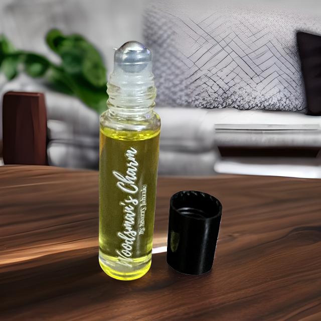 A small bottle of yellow essential oil labeled "Woodsman's Charm By Snazzy Jahzzie" with a roller applicator sits open on a wooden surface. A black cap lies next to it. In the blurred background, there is a bed with grey and white bedding and a green plant, enhancing the room's natural allure.