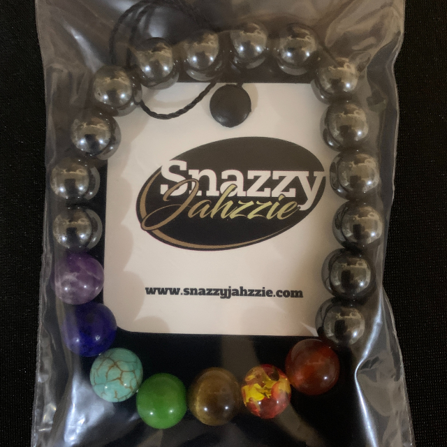 A beaded bracelet with silver and colorful Chakra beads is displayed in clear plastic packaging. The label inside reads "Snazzy Jahzzie LLC" with the website URL "www.snazzyjahzzie.com." The HEMATITE CHAKRA BRACELET features a mix of silver, purple, blue, green, and red colors for optimal energy balance.