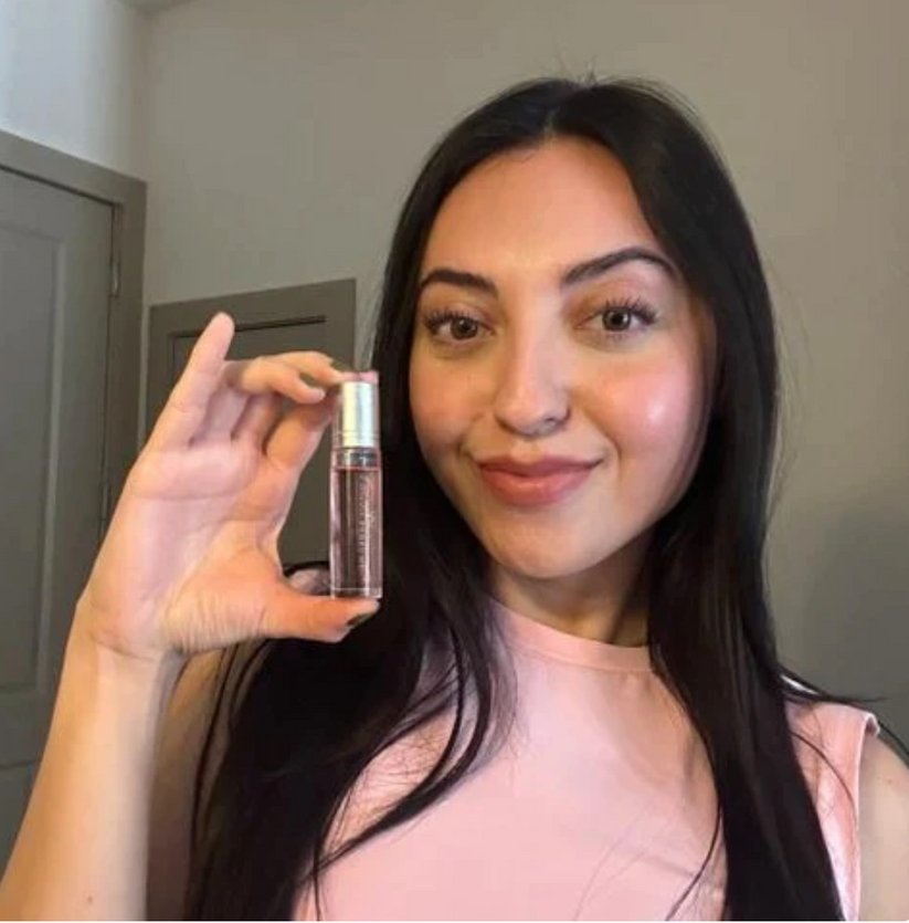 A woman with long dark hair is smiling and holding up a small bottle of clear liquid with a golden cap, revealing it's Snazzy Jahzzie® Phero Perfume by Snazzy Jahzzie LLC. She is wearing a light pink top. The background includes a door and a light-colored wall.