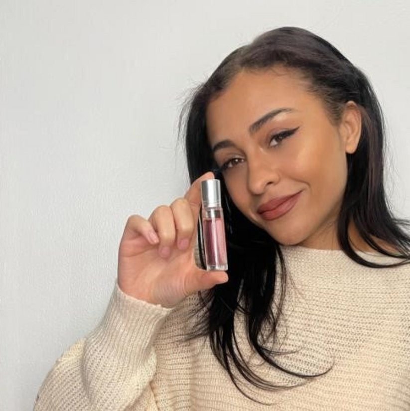 A woman with long dark hair, wearing a light beige sweater and winged eyeliner, holds up a small bottle of lipstick with a smile. The lipstick, in a transparent container with a silver cap, is reminiscent of the elegance found in Snazzy Jahzzie® Phero Perfume by Snazzy Jahzzie LLC. She stands against a plain light-colored background.