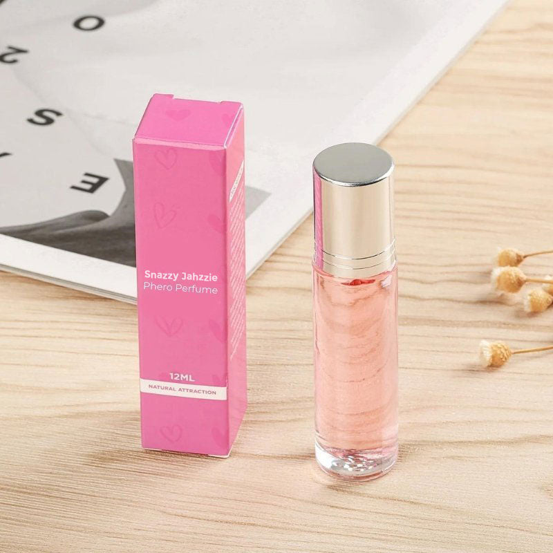 A small pink perfume bottle with a silver cap stands next to its matching rectangular pink packaging labeled "Snazzy Jahzzie® Phero Perfume, 12ml, Natural Attraction." The items are placed on a light wooden surface, with a partially visible magazine in the background.